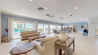 Located in Gulf Gate Estates, Sarasota, this expansive single-story community facility offers a well-rounded layout designed for gathering, recreation, and wellness. Light wood-look flooring flows throughout the main areas, complementing a bright, coastal-inspired aesthetic with soft blue and neutral tones.

The open living and dining area is bathed in natural light from large sliding glass doors that frame direct views of an outdoor pool and patio. Crown molding and recessed lighting add a polished touch, while the dining space features a charming white brick accent wall and plantation shutters. The space connects seamlessly to two fully equipped kitchens, each featuring stainless steel appliances, granite countertops, white shaker cabinetry, and tile backsplashes — ideal for catering events or community use.

A dedicated exercise room provides a comprehensive fitness experience, housing a wide array of equipment including cable machines, rowing machines, ellipticals, stationary bikes, and free weights, all set on durable commercial-grade carpet. An additional game or lounge area with a pool table adds a recreational dimension to the space.

The welcoming entryway leads through a bright corridor into the various zones of the facility. Throughout, recessed lighting, large windows, and a cohesive coastal palette create an inviting, functional environment suited for community gatherings and active lifestyles.