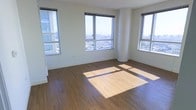 This single-floor space offers an open, airy layout defined by rich hardwood flooring and crisp white walls that lend a clean, modern aesthetic. Three large windows—positioned across multiple walls—flood the interior with abundant natural light, casting warm sunlight patterns across the floor throughout the day. The elevated position of the unit provides expansive panoramic city views, visible from multiple angles within the main living area, making the windows a true architectural highlight. A subtle cylindrical column near the center of the room adds a distinctive structural design element while preserving the open flow of the space. The generously proportioned main living area offers considerable flexibility for furniture arrangement and use. Neutral tones throughout—from the pale walls to the warm wood flooring—create a versatile backdrop suited to a variety of interior design styles. The overall ambiance is bright, modern, and urban, with the combination of elevated city views, natural light, and quality finishes contributing to a polished and inviting atmosphere.