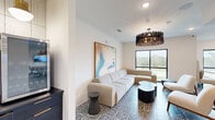 This thoughtfully designed single-floor amenity space comprises three distinct areas, each serving a unique function while maintaining a cohesive modern aesthetic. Throughout the space, an eye-catching patterned tile floor with swirling topographic motifs anchors the interiors and ties the rooms together visually.

The lounge area is a welcoming social hub, featuring a generous sectional sofa in soft white upholstery, mid-century inspired accent chairs, and sleek dark side tables. A bold, oversized abstract painting in muted blues and warm tones adorns the wall, while a striking sculptural chandelier with organic, branch-like elements adds dramatic overhead interest. Large black-framed windows invite abundant natural light and offer pleasant outdoor views.

The conference room is designed for productivity, centered around a spacious dark conference table surrounded by modern upholstered chairs. A wall-mounted display screen facilitates presentations, and a sculptural woven pendant light serves as an artistic focal point. Black-framed glass doors and windows maintain visual connectivity with the rest of the space.

A dedicated podcast and recording studio rounds out the suite, featuring a bold geometric-patterned accent wall, a glowing neon "ON AIR" sign, professional recording equipment, and a green screen curtain — making it a fully functional content creation space.