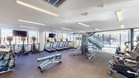 This contemporary fitness center is designed to provide a comprehensive workout experience within a bright, open environment. The space is defined by expansive floor-to-ceiling windows that wrap around much of the perimeter, flooding the interior with natural light and offering views of the surrounding urban cityscape — making cardio sessions especially engaging.

The cardio zone is prominently featured along the windowed walls and includes a row of commercial-grade treadmills, elliptical trainers, and a stationary bike, all positioned to take full advantage of the outdoor views. A wall-mounted flat-screen television adds an entertainment option for those on the machines.

The strength training area is equally well-appointed, featuring a multi-station cable and weight machine, a dedicated free weights section with a full rack of dumbbells in varying sizes, and adjustable workout benches positioned throughout the floor. The durable, rubberized flooring provides both safety and practicality across all training zones.

A glass-enclosed staircase is visible adjacent to the gym, adding an architectural element that contributes to the open, airy feel of the space. The crisp white walls and recessed linear lighting maintain a clean, modern aesthetic throughout. A separate utility and changeroom corridor is accessible from within the fitness area, offering added convenience for users.