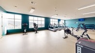 This fitness center in Madison, Wisconsin, offers a generously sized, single-floor exercise room designed to accommodate a wide range of workout routines. The space is defined by its bold teal accent walls, which contrast cleanly against a white ceiling and neutral-toned speckled flooring, creating an energetic yet functional atmosphere conducive to focused training.

Natural light fills the room through a row of large windows along one wall, providing views of the surrounding neighborhood and helping to maintain an open, airy feel throughout the space. Overhead, flush-mounted rectangular fluorescent light fixtures ensure consistent, even illumination across the entire floor area.

The exercise room is well-stocked with a comprehensive selection of equipment. A lineup of treadmills is arranged along the windowed wall, accompanied by stationary bikes and a rowing machine for cardio training. On the opposite side, a multi-tiered dumbbell rack and a weight bench provide options for strength and resistance training. Additional specialty equipment, including a plyo box and a functional training bike, rounds out the facility's offerings.

The layout is thoughtfully organized, leaving ample open floor space between equipment stations to allow for comfortable movement and stretching. A wall-mounted television and posted informational materials add practical amenities to this clean, purpose-built fitness environment.