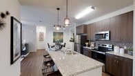 This contemporary single-floor residence is designed with an open-concept layout that seamlessly connects the kitchen, dining, and living areas, creating an inviting and functional living environment. The kitchen is a standout feature, anchored by a generous granite-topped island with a double sink and bar seating — ideal for casual dining or entertaining. Rich, dark wood cabinetry provides ample storage and contrasts beautifully with the light stone countertops, while a mosaic tile backsplash adds a refined decorative touch. The kitchen is fully equipped with stainless steel appliances, including a refrigerator, microwave, and range. Overhead, stylish industrial-style pendant lights hang above the island, adding warmth and character to the space. Durable wood-look flooring flows throughout, lending a cohesive and modern aesthetic. The adjacent dining area accommodates a full table and chairs, framed by a vibrant piece of wall art that introduces color and personality to the space. The open sightlines throughout the floor plan contribute to an airy, spacious feel, while neutral wall tones and contemporary finishes ensure a clean, polished ambiance throughout.