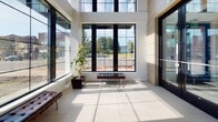 Located at 125 Bloomfield Ave in Montclair, New Jersey, this single-floor commercial lobby presents a striking first impression through its contemporary architectural design and refined finishes. The space is defined by dramatic double-height ceilings that amplify its sense of openness, complemented by expansive floor-to-ceiling grid-patterned windows that flood the interior with abundant natural light and offer clear views of the surrounding streetscape along Bloomfield Avenue.

Large-format light-colored porcelain tile flooring runs throughout, contributing to the clean, modern aesthetic. A bold marble-clad accent column serves as a visual centerpiece, rising to full height and anchoring a wall-mounted display screen. The warm, textured wood-paneled wall cladding provides a balanced contrast to the crisp white surfaces throughout the space.

The entryway features sleek black-framed glass doors that maintain the transparency and openness of the design. Thoughtfully placed seating, including tufted leather benches in a classic style, adds a welcoming touch, while tall potted tropical plants introduce organic warmth to the otherwise polished environment. A glass and metalwork accent table near the entrance completes the sophisticated furnishing palette. The overall ambiance is bright, airy, and professional, making this lobby a compelling and functional gateway for any visitor.