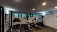 This single-level fitness studio is thoughtfully designed to support a comprehensive workout experience. The space is divided into distinct exercise zones, with cardio equipment—including treadmills, elliptical trainers, and stationary bikes—positioned along one wall beneath a row of horizontal windows that allow natural light and outdoor greenery views. Rubber floor mats are placed beneath each machine, protecting the durable carpeted flooring throughout.

The strength-training area features a multi-station cable machine alongside free weights and medicine balls in a variety of sizes, stored neatly along the wall. A large floor-to-ceiling mirror spans one wall, visually expanding the space and allowing users to monitor their form during exercise.

The studio's interior walls are painted in a calming light blue and adorned with motivational typography in varying fonts and sizes—words such as "commitment," "strength," "focus," "excellence," and "motivation" create an energizing atmosphere. White shiplap paneling on the lower portion of the walls adds a clean, modern contrast. Recessed ceiling lighting provides consistent illumination throughout. The entrance features a glass-panel door, and a water cooler is conveniently available near the entry. Overall, the space maintains a motivating, functional, and well-organized ambiance.