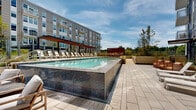 The outdoor amenity space at 440 Calvert Ave in College Park, Maryland, presents a thoughtfully designed resort-style pool deck integrated within a contemporary multi-story building complex. The centerpiece is a generously sized rectangular pool finished with deep navy blue tile along its exterior walls, giving it a sleek, modern aesthetic that contrasts beautifully with the clear, turquoise water. The pool is framed by a spacious paved deck constructed from large-format concrete pavers, providing ample room for circulation and relaxation.

A covered pergola structure with warm wood accents runs along one side of the deck, sheltering a row of white lounge chairs ideal for sunbathing or leisurely outdoor gatherings. On the opposite side of the pool, clusters of contemporary lounge seating in warm tones offer additional gathering areas. Ornamental grasses and carefully curated plantings are integrated throughout raised planter beds, introducing natural texture and greenery that soften the hardscape environment.

The deck is accessible via paved walkways with stainless steel railing systems, ensuring safe and convenient circulation throughout the space. Floor-to-ceiling glass walls of an adjacent interior amenity room open visually onto the pool area, reinforcing a seamless indoor-outdoor connection. The surrounding multi-story building facades, clad in contrasting light and dark panel systems with expansive windows, frame the space and provide a striking architectural backdrop under open skies.