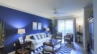 The living room serves as the centerpiece of this single-floor space, immediately drawing attention with its dramatic royal blue accent wall that adds depth and personality to the interior. Crown molding frames the room's perimeter, lending an elegant architectural touch that complements the bold color scheme. A ceiling fan is centrally mounted, offering both practical airflow and a clean visual anchor for the room.

Natural light flows generously through a sliding glass door fitted with vertical blinds and patterned drapes, providing a view of an outdoor area and brightening the space throughout the day. The living area is furnished with a classic cream sofa adorned with decorative pillows, paired with dark wood accent chairs featuring patterned upholstery. A substantial dark wood coffee table anchors the seating arrangement over a bold navy and white chevron area rug, reinforcing the room's cohesive color palette.

Additional furnishings include side tables with table lamps that provide warm ambient lighting. A tall indoor plant near the sliding door introduces a natural element, softening the room's overall aesthetic while complementing its rich, curated design.