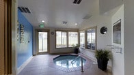 This single-floor indoor spa facility centers around a recessed octagonal hot tub, elegantly set into a tiled surround and accented with a stainless steel entry rail. The room is finished with light-toned tile flooring throughout, creating a clean and functional aesthetic well-suited to a relaxation space. Large windows line the walls, flooding the interior with natural light and offering views of an adjacent outdoor patio and landscaped surroundings. A glass-paneled door provides direct access to the exterior, enhancing the connection between the indoor and outdoor environments. Recessed ceiling lighting supplements the natural light, ensuring the space is well-illuminated at all times. Potted tropical plants are positioned throughout the room, adding a touch of greenery and a welcoming, resort-like ambiance. The walls are finished in a neutral palette with a subtle blue accent wall, contributing to the calm, serene atmosphere. Ventilation is handled by ceiling-mounted HVAC vents, maintaining a comfortable environment year-round. This space is well-appointed for relaxation and leisure, combining practical amenities with an inviting, light-filled atmosphere.