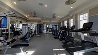 Located at 722 Island Park Dr. on Daniel Island, SC, this single-floor fitness center offers a comprehensive workout environment designed to accommodate a wide range of exercise needs. The space is outfitted with a robust selection of cardio equipment, including ellipticals, stationary bikes, and treadmills, arranged in orderly rows to maximize usability and flow. Strength training machines line the perimeter, providing users with a full suite of resistance equipment. Large windows along the walls invite generous natural light, creating an open and energizing atmosphere. The interior features a tray ceiling accented with crown molding, lending an elevated architectural detail uncommon in typical fitness facilities. Multiple ceiling fans are distributed throughout, ensuring consistent air circulation. Recessed lighting supplements the natural light for well-illuminated workouts at any time of day. Full-length mirrors along one wall expand the visual depth of the room while allowing users to monitor their form. The durable rubber flooring provides both safety and comfort underfoot. A mounted television is positioned for easy viewing from multiple workout stations, enhancing the overall experience.