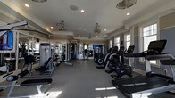 Located at 722 Island Park Dr. on Daniel Island, SC, this single-floor fitness center offers a comprehensive workout environment designed to accommodate a wide range of exercise needs. The space is outfitted with a robust selection of cardio equipment, including ellipticals, stationary bikes, and treadmills, arranged in orderly rows to maximize usability and flow. Strength training machines line the perimeter, providing users with a full suite of resistance equipment. Large windows along the walls invite generous natural light, creating an open and energizing atmosphere. The interior features a tray ceiling accented with crown molding, lending an elevated architectural detail uncommon in typical fitness facilities. Multiple ceiling fans are distributed throughout, ensuring consistent air circulation. Recessed lighting supplements the natural light for well-illuminated workouts at any time of day. Full-length mirrors along one wall expand the visual depth of the room while allowing users to monitor their form. The durable rubber flooring provides both safety and comfort underfoot. A mounted television is positioned for easy viewing from multiple workout stations, enhancing the overall experience.