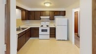 This single-floor residential unit offers a practical and well-organized layout across several distinct living spaces. The living room is generously proportioned, featuring light neutral carpeting, a wall-mounted air conditioning unit, baseboard heating, and a large sliding glass door flanked by vertical blinds that allow abundant natural light to fill the room. A pass-through window connects the living area to the kitchen, enhancing the sense of openness between spaces. The kitchen is efficiently designed with rich dark wood cabinetry extending to the upper walls, light countertops, a tile backsplash, and white appliances including a refrigerator and electric range. Vinyl flooring and a ceiling light fixture complete the functional kitchen space. Adjacent to the living room, the dining area is distinguished by a classic chandelier overhead and warm carpet underfoot, making it a defined and inviting space for meals. The bedroom features soft carpeting, a window with blinds offering outdoor views and natural light, textured ceilings, and baseboard heating. Throughout the unit, warm wood-toned baseboards provide consistent visual continuity, and white walls keep the interiors feeling bright and clean. The overall layout, as seen in the dollhouse view, is compact yet thoughtfully arranged for comfortable everyday living.