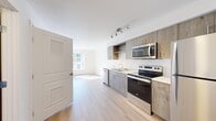 Located at 13360 SE Eastgate Way in Bellevue, WA, this two-bedroom, one-bathroom unit offers a thoughtfully designed single-floor layout that maximizes every square foot of its compact footprint. The open kitchen anchors the living space and showcases wood-toned cabinetry with a weathered finish, stainless steel appliances including a refrigerator, electric range, built-in microwave, and dishwasher, and light-toned countertops. Track lighting overhead provides focused illumination throughout the kitchen area. Light hardwood-style flooring runs through the main living and kitchen areas, contributing to a bright, clean aesthetic. The bedrooms are carpeted for added comfort, with the primary bedroom featuring a ceiling fan, a window that allows natural light, and a walk-in closet equipped with wire shelving. The bathroom is finished with wood-toned cabinetry, a single-basin vanity, a full soaking tub with a curved shower rod, built-in wall shelving, and durable hardwood-style flooring. White walls and neutral finishes throughout create a cohesive, modern ambiance, while the efficient layout ensures each room remains functional and well-defined.