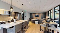 This single-floor community amenity space is thoughtfully designed to serve as a stylish gathering hub for residents. The open-concept layout seamlessly connects a fully equipped kitchen, a dining area, and a lounge zone, creating a cohesive and functional environment throughout.

The kitchen is outfitted with stainless steel appliances — including a full-size refrigerator, gas range, and dishwasher — complemented by dark cabinetry, a tile backsplash, and a central island with bar seating and pendant lighting above. Light hardwood flooring runs throughout the space, lending warmth and continuity.

The dining area is anchored by a rectangular table with upholstered seating for six, positioned beneath a pair of statement pendant lights. Floor-to-ceiling windows flood the area with natural light and offer direct views of an outdoor courtyard and terrace.

The lounge area features plush sofas arranged around a low-profile coffee table, all oriented toward a large flat-screen TV mounted on a warm wood accent wall set against a deep charcoal backdrop. A casual high-top table and bar seating provide additional social gathering space. Large black-framed windows throughout the lounge further connect the interior to the outdoor surroundings. The overall ambiance is contemporary and inviting, balancing comfort with a clean, modern aesthetic.