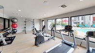 This fitness center is designed to support a comprehensive workout experience within a clean, modern space. The layout is open and well-organized, with patterned carpet flooring throughout that adds a contemporary aesthetic while providing comfort underfoot. Recessed lighting illuminates the entire area evenly, creating a bright and energizing atmosphere.

The cardio zone is generously equipped with multiple treadmills, elliptical trainers, and stationary bikes, offering ample options for aerobic exercise. A cable machine and a full power rack with weight plates and barbells anchor the strength training section, complemented by a dedicated set of stability balls in varying sizes for functional training. The dumbbell rack and additional free weights round out the strength offerings.

Large black-framed windows line the walls, flooding the space with natural light and offering views of the outdoor pool area complete with lounge chairs and umbrellas — a motivating backdrop for any workout. An interior window connects the fitness center to an adjacent lounge or common area, enhancing the sense of openness. A glass door provides direct access to the outdoor pool deck, making it easy to transition between indoor and outdoor amenities. The overall ambiance is modern, clean, and purpose-built for an active lifestyle.