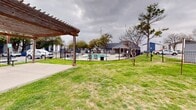 This outdoor community amenity space at 9200 N Normandale St in Fort Worth, Texas, offers a well-maintained gathering area designed for relaxation and recreation. The centerpiece of the space is a sturdy, dark-stained wood pergola structure, supported by four posts and covering a concrete patio pad. The pergola provides a shaded seating area and includes a picnic table beneath it, making it a practical spot for outdoor dining or casual gatherings. Surrounding the pergola is a generous expanse of open green lawn, providing ample room for outdoor activities. The grounds are enclosed by a black iron fence that runs the perimeter of the space, offering both security and a defined boundary. A standout feature visible from the lawn area is a fenced swimming pool, finished in a classic blue tone and surrounded by additional iron fencing for safety. Mature trees are scattered throughout the property, offering natural shade and adding to the greenery of the outdoor environment. The residential buildings of the surrounding community are visible in the background, providing context to the communal nature of this outdoor space. Overall, this amenity area is a functional and accessible outdoor retreat within a residential community setting in Fort Worth.