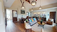 This well-appointed single-floor space showcases a cohesive nautical-inspired design blended with warm, rustic elements throughout. The main living area commands attention with soaring vaulted ceilings and a floor-to-ceiling stacked stone fireplace, complete with a richly stained wood mantel and a wall-mounted television above. Large windows flank the fireplace, flooding the room with natural light and providing views of the surrounding greenery. Pendant and drum-style light fixtures hang from the high ceilings, adding character and warmth to the space. Rich, wide-plank hardwood flooring runs throughout, unifying each area seamlessly. The dining area features woven rattan chairs and dark wood tables, accented by classic board-and-batten wainscoting along the walls. Decorative crossed oars overhead reinforce the nautical theme, as do maritime artwork and compass motifs displayed throughout. Dark-stained glass-panel interior doors add an elegant, crafted detail as they connect the various areas of the space. The kitchen is outfitted with warm natural wood cabinetry, granite countertops, stainless steel appliances, and stone tile accents. A dedicated bar or service counter area provides additional functionality. Overall, the space delivers a comfortable, inviting ambiance with a strong sense of craftsmanship and thematic cohesion.
