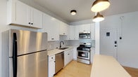 Located at 1033 Hyperion Ave in the vibrant Silver Lake neighborhood of Los Angeles, this single-floor space showcases a clean, modern aesthetic with carefully selected finishes throughout. The kitchen is a standout feature, outfitted with crisp white shaker cabinets accented by matte black hardware, a geometric hexagonal tile backsplash, and a full suite of stainless steel appliances including a gas range, over-the-range microwave, dishwasher, and refrigerator. Light wood flooring and pendant lighting lend warmth to the kitchen's contemporary feel, while a breakfast bar provides additional functionality. The living area continues the light wood flooring and is topped by a sleek black ceiling fan, creating a bright and airy atmosphere. An open doorway leads to a walk-in closet or additional storage space, adding practicality to the layout. The bathroom features floor-to-ceiling white subway tile in a vertical stacked pattern within the tub and shower enclosure, complemented by matte black fixtures for a cohesive, modern touch. A window allows natural light to fill the space. Overall, the unit presents a well-curated, move-in-ready environment with a consistent design language of white tones, natural wood, and black accents.