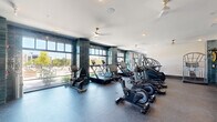 Located at 1045 Williamsburg Way in Evans, Georgia, this fitness center offers a well-appointed, single-floor layout designed to support a comprehensive workout experience. The space is divided into two distinct functional zones: a cardio area and a strength training room.

The cardio zone is the standout area of the facility, featuring floor-to-ceiling windows framed by striking teal glazed tile columns that flood the room with natural light and provide open views of the outdoors. True-brand upright and recumbent bikes, treadmills, ellipticals, and stair climbers are arranged to maximize both usability and the view. Ceiling fans with brass accents add both style and airflow throughout.

The strength training room is a well-lit, quieter space equipped with a TKO multi-station cable machine, a weight bench, free weights, and additional resistance equipment. Large windows allow natural light to fill the room, creating an energizing environment for resistance training.

The entry corridor serves as a stylish transition into the fitness areas, featuring bold geometric wall art, open grid shelving, marble-topped cabinetry with brass hardware, and a small under-counter refrigerator — providing a convenient refreshment station for users. The durable rubber flooring throughout the cardio and strength areas ensures safety and comfort during workouts.
