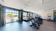 Located at 1045 Williamsburg Way in Evans, Georgia, this fitness center offers a well-appointed, single-floor layout designed to support a comprehensive workout experience. The space is divided into two distinct functional zones: a cardio area and a strength training room.

The cardio zone is the standout area of the facility, featuring floor-to-ceiling windows framed by striking teal glazed tile columns that flood the room with natural light and provide open views of the outdoors. True-brand upright and recumbent bikes, treadmills, ellipticals, and stair climbers are arranged to maximize both usability and the view. Ceiling fans with brass accents add both style and airflow throughout.

The strength training room is a well-lit, quieter space equipped with a TKO multi-station cable machine, a weight bench, free weights, and additional resistance equipment. Large windows allow natural light to fill the room, creating an energizing environment for resistance training.

The entry corridor serves as a stylish transition into the fitness areas, featuring bold geometric wall art, open grid shelving, marble-topped cabinetry with brass hardware, and a small under-counter refrigerator — providing a convenient refreshment station for users. The durable rubber flooring throughout the cardio and strength areas ensures safety and comfort during workouts.
