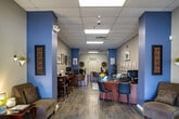 Located at 1501 Little Gloucester Road in Blackwood, NJ, this single-floor commercial office suite offers a thoughtfully designed, open-plan workspace that balances professionalism with a welcoming atmosphere. The space is anchored by a bright, glass-door entrance that floods the foyer with natural light and offers views of the surrounding parking area and landscaping. A branded entry mat greets visitors as they step inside.

The interior is defined by a neutral and blue-toned color palette, with accent columns and walls in slate blue contrasting against light gray and white surfaces. Wood-look plank flooring runs throughout, adding warmth and cohesion to the entire space. A drop ceiling with recessed panel lighting ensures consistent, well-distributed illumination across all areas.

Toward the front, a comfortable lounge area features upholstered seating, a patterned area rug, and a display wall showcasing property floor plans and renderings — indicative of a real estate or sales office environment. A central reception desk with client-facing seating provides a clear focal point for staff-client interaction. Further into the space, additional workstations and a conference-style table support collaborative work. Framed artwork and decorative accents are placed throughout, contributing to a polished yet approachable ambiance.