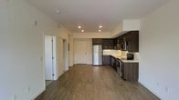 This single-floor unit offers a thoughtfully designed open-concept layout that seamlessly integrates the living and kitchen areas into one cohesive space. The kitchen is a standout feature, equipped with dark espresso-toned cabinetry that extends to the upper and lower sections of the walls, providing generous storage. Stainless steel appliances — including a refrigerator, range, microwave, and sink — lend a contemporary, functional appeal. Light-toned countertops offer a clean contrast against the darker cabinetry, enhancing the modern aesthetic.

The flooring throughout the space consists of warm, medium-toned hardwood-style planks that add continuity and a welcoming feel. Recessed lighting is strategically placed across the ceiling, ensuring consistent, even illumination across the open area. The neutral white walls and ceilings contribute to a bright and airy atmosphere.

Additional doorways visible from the main living area suggest access to separate rooms or storage spaces, adding to the functional versatility of the layout. The overall design is clean, modern, and practical.