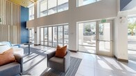 Located at 5805 Charlotte Dr in San Jose, CA, this generously sized resident lounge is a contemporary communal space designed to foster comfort and community. The single-floor layout spans a substantial open footprint, showcasing soaring double-height ceilings that create a grand, airy atmosphere throughout.

The space is anchored by a striking central lobby area featuring bold navy blue accent walls, warm wood-paneled ceilings, and a mix of curved modular sofas in neutral tones accented with vibrant orange cushions. A full-sized indoor tree serves as a natural focal point, adding life and warmth to the interior. Circular porthole-style windows at the upper mezzanine level add an architectural detail that enhances the visual depth of the space.

Floor-to-ceiling sliding glass doors line one side of the lounge, flooding the interior with natural light and providing seamless access to an outdoor area framed by palm trees. Large-format tile flooring and patterned carpet zones delineate distinct seating and activity areas throughout.

A dedicated dining and kitchen area features a long communal wood dining table, modern dark cabinetry, built-in appliances, and mounted displays — ideal for gatherings or casual use. Additional lounge zones offer curved blue sectional seating, bistro-style tables, and a sculptural pendant ceiling fixture, completing this well-appointed and versatile community space.