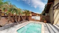 This single-level outdoor community space offers a well-appointed and relaxing environment designed for leisure and social gathering. The centerpiece of the space is a generously sized spa and hot tub, set within a tiled surround and enclosed by a classic brick perimeter wall. Tall, mature palm trees rise alongside the wall, lending a resort-like ambiance and a sense of established, tropical character to the setting. The warm, sunny climate is reflected throughout the design, with open skies above and abundant natural light bathing the entire area.

Adjacent to the spa, a spacious covered patio provides a comfortable outdoor dining and lounge area. A sturdy wood-beam pergola overhead casts striking striped shadows across the concrete floor, creating a visually dynamic and partially shaded retreat. Multiple round metal dining tables, each surrounded by matching lattice-style chairs, are arranged throughout the patio, accommodating groups of varying sizes. The pergola structure connects seamlessly to the surrounding walls, providing partial shelter while maintaining an open, airy feel.

The overall aesthetic is warm and practical, with a Southern California character evident in the brick walls, palm landscaping, and sun-drenched hardscape. This outdoor space is well-suited for communal relaxation and casual outdoor dining.