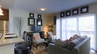 This single-floor living space presents a well-appointed interior that blends contemporary style with a warm, inviting ambiance. The living area serves as the focal point of the space, anchored by a large window dressed with vertical blinds that allows abundant natural light to fill the room. The window also provides a view of an outdoor area, suggesting balcony or patio access.

The room features a thoughtfully curated design scheme built around a dark and neutral color palette, accented with bold pops of orange and red through decorative pillows and patterned upholstery. A plush sectional sofa and a pair of accent chairs create a comfortable and functional seating arrangement centered over a colorful area rug that adds texture and visual interest to the floor.

Along one wall, a dark media console houses a flat-screen television and is flanked by a coordinating dresser. Above, an arrangement of black-framed mirrors of varying sizes adds depth and a decorative focal point to the wall. Soft ambient lighting is provided by a floor lamp. The overall atmosphere is modern and cohesive, reflecting a carefully considered interior design aesthetic.