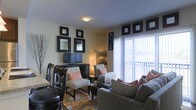 This single-floor living space presents a well-appointed interior that blends contemporary style with a warm, inviting ambiance. The living area serves as the focal point of the space, anchored by a large window dressed with vertical blinds that allows abundant natural light to fill the room. The window also provides a view of an outdoor area, suggesting balcony or patio access.

The room features a thoughtfully curated design scheme built around a dark and neutral color palette, accented with bold pops of orange and red through decorative pillows and patterned upholstery. A plush sectional sofa and a pair of accent chairs create a comfortable and functional seating arrangement centered over a colorful area rug that adds texture and visual interest to the floor.

Along one wall, a dark media console houses a flat-screen television and is flanked by a coordinating dresser. Above, an arrangement of black-framed mirrors of varying sizes adds depth and a decorative focal point to the wall. Soft ambient lighting is provided by a floor lamp. The overall atmosphere is modern and cohesive, reflecting a carefully considered interior design aesthetic.
