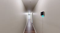 This thoughtfully laid-out single-floor unit offers a functional and versatile living arrangement across five distinct rooms. A narrow hallway with dark hardwood-style flooring and neutral walls connects the spaces, with a wall-mounted smart device adding a modern touch at the entrance.

The office is a standout feature, configured as a professional workspace with a round conference table, ergonomic chairs, a sleek black futon sofa, and tall potted plants that add a touch of greenery. Natural light filters in through slatted blinds, creating a bright and productive environment.

The bedroom is a comfortable and well-lit retreat, featuring a window with blinds, warm hardwood-style flooring, and enough space for a full-sized bed along with a cozy seating area equipped with a cushioned chair, side table, and decorative plants.

The kitchen is efficiently designed with grey cabinetry, dark granite countertops, a stainless steel range, a black dishwasher, and a full-sized refrigerator. A dining area with a wooden table and window provides a pleasant spot for meals.

The bathroom is clean and modern, featuring grey tile surrounding a soaking tub with shower, a white vanity with marble-style countertop, and tile flooring. Consistent neutral tones and hardwood-style floors throughout the unit create a cohesive, understated aesthetic.