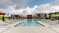 The pool amenity area at The Waters in Hammond, Louisiana, delivers a thoughtfully designed outdoor retreat centered around a large, rectangular swimming pool with calm, clear water. The expansive concrete pool deck surrounds the pool on all sides, accommodating numerous lounge chairs and green accent seating, providing ample space for relaxation under the open Louisiana sky. A striking wood-slatted cabana structure serves as a focal point at the pool's edge, adding architectural character to the setting.

Adjacent to the pool, a covered outdoor lounge pavilion features exposed wood beam construction, ceiling fans, and a brick fireplace wall mounted with an outdoor television—ideal for year-round gatherings. Comfortable wicker and upholstered seating arrangements make this shaded area a natural extension of the pool deck. Large glass windows and doors connect the pavilion to an interior fitness area, blending indoor and outdoor amenities seamlessly.

The entry to the clubhouse is framed by exposed brick columns and floor-to-ceiling black-framed glass doors, offering views directly through to the pool beyond. Manicured landscaping with ornamental plants, decorative mulch beds, and accent lighting borders the pathways, enhancing the polished, resort-inspired atmosphere throughout the community.