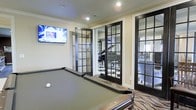 Located at 5315 Avery Road in Dublin, Ohio, this single-floor amenity space is designed to offer residents a refined and social environment. The billiards room serves as a central highlight, featuring a full-sized pool table set atop a patterned area rug, recessed lighting, and crown molding that adds a polished architectural touch. A wall-mounted flat-screen television enhances the entertainment experience in this area. Striking black-framed French doors with multi-pane glass panels connect the billiards room to adjacent spaces, allowing natural light to flow through while maintaining a sense of openness and visual continuity. Through these doors, a grand piano is visible in a neighboring lounge, suggesting a dedicated music or social room ideal for gatherings and events. Beyond that, an additional room — likely a lounge or dining area — can be glimpsed, hinting at a well-appointed layout designed for community use. The overall aesthetic blends classic architectural detailing with contemporary finishes, creating an inviting and upscale atmosphere throughout. The space is well-suited for leisure, entertainment, and social interaction.
