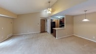 This single-floor residence presents a well-organized layout with a cohesive neutral color palette of warm beige tones carried throughout. The open-concept living area is a standout feature, showcasing vaulted ceilings accented by a ceiling fan with light fixture, creating an airy and spacious atmosphere. Plush wall-to-wall carpeting extends across the living space, lending warmth underfoot. A peninsula-style kitchen overlooks the living area and is equipped with rich dark wood cabinetry, laminate countertops, a stainless steel sink, and black appliances including a refrigerator, electric range, and microwave. A closet near the entry provides additional storage, and in-unit laundry is conveniently located nearby.

The bedrooms are generously proportioned, each featuring soft carpeting, neutral walls, and windows fitted with horizontal blinds that allow natural light to filter in. One bedroom includes a walk-in closet with built-in shelving. The unit includes two bathrooms: a full bathroom with a tub/shower combination, a large vanity mirror, dark wood cabinetry, laminate countertops, and wood-look flooring; and a half bathroom with a single-sink vanity and matching wood-look flooring. Overall, the space presents a functional and comfortable layout with consistent finishes throughout.