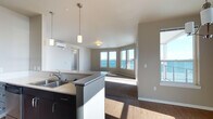 This single-floor space offers a thoughtfully designed open-concept layout that seamlessly connects the kitchen and living areas. The kitchen is anchored by a dark-cabinetry island with a built-in double sink and a stainless steel countertop, complemented by stylish pendant lighting that adds a modern touch overhead. A wall-mounted mini-split unit ensures year-round climate comfort.

The transition from kitchen to living area flows naturally across durable hardwood-style flooring, which gives way to plush carpeting in the main living space. A curved bay of large windows wraps around the living area, flooding the interior with abundant natural light and framing expansive panoramic views of what appears to be open water and the surrounding skyline. These windows create a striking focal point and a strong sense of openness throughout the space.

The neutral color palette — featuring soft gray and warm beige tones on the walls — enhances the clean, contemporary aesthetic. Additional windows along the side walls further amplify the natural brightness and ventilation. The overall ambiance is airy, light-filled, and modern, making this space well-suited for those who appreciate open layouts, scenic views, and a minimalist yet functional design.