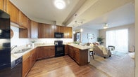 This single-floor residence showcases an open-concept layout that connects the kitchen and living areas in a cohesive, functional design. The kitchen is a standout feature, outfitted with rich, warm-toned wood cabinetry that extends to the ceiling, offering generous storage throughout. Granite countertops in soft neutral tones provide ample workspace, complementing the black appliance suite that includes an electric range, built-in microwave, and additional appliances. A peninsula with seating extends from the main counter, creating a natural transition between the kitchen and living space while offering a casual dining option.

Hardwood flooring runs through the kitchen area, adding warmth and visual continuity, while transitioning to carpet in the living room. Recessed and pendant lighting illuminate the kitchen, with a track light fixture adding a modern touch. The adjacent living area benefits from natural light streaming through large windows, creating a bright and welcoming ambiance. A ceiling fan enhances comfort in the living space, which features neutral wall tones that allow for versatile décor. The overall layout is practical and inviting, with a smooth flow between functional zones.