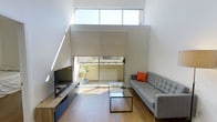 This well-appointed 2-bedroom, 2-bathroom residence offers a thoughtfully designed living environment with standout architectural details. The living area immediately impresses with its dramatic double-height ceilings, which flood the space with natural light through oversized upper clerestory windows and sliding glass doors that open to an outdoor patio area. The open, airy atmosphere is enhanced by clean white walls and warm hardwood flooring that runs throughout.

The living room is anchored by a sleek media console and a modern glass coffee table, complemented by a contemporary gray sofa and direct access to a private outdoor space with greenery. The sliding glass door and large windows work together to seamlessly blur the boundary between interior and exterior, creating a bright and inviting atmosphere.

The minimalist design aesthetic is consistent across the home, with neutral tones and clean lines defining each room. The two bedrooms provide comfortable private retreats, while the two full bathrooms offer functional convenience. The overall layout makes efficient and intelligent use of the available space, balancing openness in the common areas with well-defined private zones. This residence is ideal for those who appreciate modern architecture, natural light, and a clean, contemporary living environment.