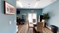 This single-floor space is designed to serve as a comfortable and functional communal gathering area. The interior is defined by warm teal-painted walls that lend a calm, inviting atmosphere throughout. The space is organized to accommodate both dining and lounge activities within an open layout, making it versatile for a variety of social uses.

The dining area features a round glass-top table surrounded by cushioned upholstered chairs, providing a comfortable setting for meals or group gatherings. Adjacent to the dining area, a lounge zone is furnished with a classic wooden console table, decorative accents, and a wall-mounted flat-screen television, creating a relaxed seating environment. A full-size refrigerator is also present, adding practical utility to the space.

Natural light enters generously through a set of French doors fitted with white blinds, connecting the interior to the outdoors and enhancing the overall sense of openness. The patterned carpet flooring adds a layer of texture and visual interest underfoot, complementing the teal wall color. Overhead, flush-mounted fluorescent lighting ensures the space remains bright and well-lit throughout the day. Decorative artwork and potted greenery contribute finishing touches that make the environment feel curated and welcoming.