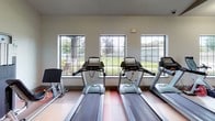 Located in Sugar Land, Texas, this well-appointed fitness center offers a comprehensive workout facility designed to accommodate a wide range of exercise needs. The space is organized into distinct zones, each tailored to a specific type of training.

The cardio area features a row of treadmills positioned in front of a bank of large, grid-paned windows that flood the space with natural light and offer pleasant views of the surrounding greenery and grounds. The bright, airy atmosphere in this section makes for an engaging workout environment.

Adjacent to the cardio zone, a dedicated cycling and functional training area houses multiple spin bikes arranged on hardwood-style flooring. Full-length mirrored walls line this section, visually expanding the space while allowing users to monitor their form. The mirrors also reflect recessed ceiling lighting, keeping the area well-lit throughout.

The strength training zone is equipped with free weights, cable machines, weight racks, and benches, alongside punching bags for combat fitness. A colorful checkerboard rubber floor anchors this section, providing both durability and traction. Soaring ceilings throughout the facility contribute to an open, spacious feel.

Glass partitions and doors divide the zones while maintaining visual connectivity across the entire floor. A wall-mounted television and additional amenity stations round out the facility, making this fitness center a fully functional and inviting space for residents.