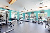 Located at 19757 Crystal Rock Dr in Germantown, MD, this well-equipped fitness center offers a single-floor layout designed to accommodate a comprehensive range of workout activities. The space is bright and energizing, characterized by bold teal accent walls, white crown molding, and an abundance of large windows that flood the interior with natural light and provide views of the surrounding greenery. Ceiling fans are strategically placed throughout to maintain airflow and comfort during workouts. The durable rubber flooring provides a safe, impact-absorbing surface across the entire exercise area. On the cardio side, multiple treadmills and elliptical machines are lined along the windowed walls, each equipped with individual monitors, with wall-mounted televisions overhead for added entertainment. The strength training area features cable machine systems, adjustable benches, and a full rack of dumbbells, complemented by large mirrors that enhance the sense of space. A dedicated functional training zone includes stability balls, medicine balls, kettlebells, and additional resistance equipment organized on a multi-tier storage rack. Recessed wood-trimmed light fixtures supplement the abundant natural light, creating a well-lit and motivating environment suitable for a wide variety of fitness routines.