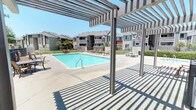 This outdoor recreational area centers around a generously sized swimming pool with clear blue water, framed by a wide concrete pool deck designed for comfortable lounging and socializing. A prominent pergola structure extends over a portion of the deck, casting distinctive striped shadows and providing a shaded retreat from the sun — ideal for relaxing without direct exposure to the elements. Multiple lounge chairs and seating areas are positioned throughout the deck, offering plenty of space for residents to unwind poolside. The area is enclosed by a secure metal fence perimeter, with a latticed security gate providing controlled access to the pool area, enhancing both safety and privacy. Adjacent to the entry, a pool rules and safety notice board is prominently displayed alongside clearly marked restroom facilities, including a dedicated men's room, ensuring convenience for users. Mature trees line the background, contributing a touch of greenery and natural shade. The surrounding multi-story residential buildings with balconies are visible beyond the fence, providing context for the community-oriented nature of this amenity space. The overall atmosphere is open, bright, and inviting, well-suited for outdoor recreation and relaxation under sunny skies.