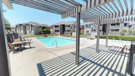This outdoor recreational area centers around a generously sized swimming pool with clear blue water, framed by a wide concrete pool deck designed for comfortable lounging and socializing. A prominent pergola structure extends over a portion of the deck, casting distinctive striped shadows and providing a shaded retreat from the sun — ideal for relaxing without direct exposure to the elements. Multiple lounge chairs and seating areas are positioned throughout the deck, offering plenty of space for residents to unwind poolside. The area is enclosed by a secure metal fence perimeter, with a latticed security gate providing controlled access to the pool area, enhancing both safety and privacy. Adjacent to the entry, a pool rules and safety notice board is prominently displayed alongside clearly marked restroom facilities, including a dedicated men's room, ensuring convenience for users. Mature trees line the background, contributing a touch of greenery and natural shade. The surrounding multi-story residential buildings with balconies are visible beyond the fence, providing context for the community-oriented nature of this amenity space. The overall atmosphere is open, bright, and inviting, well-suited for outdoor recreation and relaxation under sunny skies.