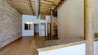 This single-floor space showcases a compelling blend of industrial and rustic design, highlighted by soaring ceilings with exposed wooden beams and visible ductwork that lend the interior a warehouse-loft character. Original timber posts punctuate the open floor plan, adding structural authenticity and visual warmth throughout.

The open-concept layout connects the kitchen and living areas seamlessly. The kitchen features granite countertops with warm tonal variation, light wood cabinetry, and ample workspace, making it both functional and visually appealing. The expansive open living area is finished with rich hardwood flooring that runs throughout, complementing the natural wood tones of the overhead beams.

A stacked washer and dryer unit is conveniently integrated into the layout, visible through one of the doorways. An elegant chandelier adds a refined touch that contrasts tastefully with the raw industrial ceiling above. Original brick detailing is visible along one wall near a framed window, reinforcing the building's historic character. The overall ambiance is open, airy, and rich in architectural detail, offering a distinctive living environment that balances character with practicality.