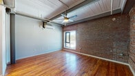 Located at 451 Pacific Cir in Mount Airy, North Carolina, this single-floor unit blends industrial character with modern conveniences. Exposed red brick accent walls serve as a defining architectural feature throughout, complemented by warm, richly toned hardwood floors that run across the entire space. The main living area is open and airy, featuring a white-painted wood plank ceiling with exposed steel beams and a ceiling fan, creating a loft-like aesthetic. A wall-mounted mini-split HVAC unit ensures year-round comfort, while a window overlooking the exterior fills the room with natural light. The galley-style kitchen is well-appointed with dark espresso cabinetry, granite-style countertops, a double stainless steel sink, and a full suite of stainless steel appliances including a refrigerator, range, and built-in microwave. A secondary room with its own brick wall and a white paneled door offers flexible use as a bedroom or additional living space. The full bathroom includes a combined tub and shower unit, a toilet, and hardwood flooring, maintaining the cohesive material palette found throughout. A generously sized walk-in closet with wire shelving and hardwood floors provides ample storage. The overall ambiance is a thoughtful mix of raw, industrial charm and comfortable, updated finishes.