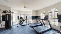 This fitness center is a thoughtfully designed, single-floor facility that offers a comprehensive workout environment for its users. The main exercise area is anchored by a generous selection of cardio equipment, including multiple treadmills, an elliptical trainer, and a stair climber, all arranged to take full advantage of the natural light that pours in through a series of large, arched windows. These elegant windows line the perimeter walls, offering views of surrounding greenery and lending the space a bright, airy atmosphere.

The walls feature a distinctive two-tone design, with a deep teal accent band contrasting against crisp white upper walls, adding visual character to the room. Blue patterned carpeting covers the floor throughout, providing cushioning underfoot. Strength training is well represented with a multi-station cable machine and additional resistance equipment, complemented by wall-mounted mirrors that enhance the sense of space and allow users to monitor their form. A ceiling fan keeps the air circulating in the primary workout zone.

A water dispenser is conveniently accessible near an arched architectural niche, and the fitness center connects directly to what appears to be a community leasing office or clubhouse area, visible through a large interior window and an open doorway. Recessed lighting and a clean, organized layout round out this well-maintained and functional fitness facility.