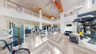 This expansive fitness center is designed with both functionality and aesthetics in mind, offering a bright, open environment for a full range of workout activities. The space is anchored by dramatic double-height ceilings accented with bold orange architectural beams, creating a visually striking interior that is further enhanced by an open mezzanine level with glass railings overlooking the main floor below.

The flooring throughout is a light-toned herringbone-patterned hardwood, lending a refined, contemporary character to the space. Floor-to-ceiling steel-framed windows along multiple walls flood the interior with abundant natural light and provide views of lush outdoor greenery, contributing to an energizing atmosphere.

The main workout area is well-equipped with a wide variety of strength machines, free weights, cable systems, and functional training equipment including TRX suspension trainers, plyo boxes, and a wall-mounted rig. A dedicated cardio zone features stationary bikes, a rower, and additional cardio machines. Large mirrors and wall-mounted televisions are positioned throughout. A built-in storage alcove with shelving and a small refrigerator adds practical convenience. The space is accessed through sleek black-framed double glass doors.