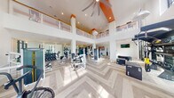 This expansive fitness center is designed with both functionality and aesthetics in mind, offering a bright, open environment for a full range of workout activities. The space is anchored by dramatic double-height ceilings accented with bold orange architectural beams, creating a visually striking interior that is further enhanced by an open mezzanine level with glass railings overlooking the main floor below.

The flooring throughout is a light-toned herringbone-patterned hardwood, lending a refined, contemporary character to the space. Floor-to-ceiling steel-framed windows along multiple walls flood the interior with abundant natural light and provide views of lush outdoor greenery, contributing to an energizing atmosphere.

The main workout area is well-equipped with a wide variety of strength machines, free weights, cable systems, and functional training equipment including TRX suspension trainers, plyo boxes, and a wall-mounted rig. A dedicated cardio zone features stationary bikes, a rower, and additional cardio machines. Large mirrors and wall-mounted televisions are positioned throughout. A built-in storage alcove with shelving and a small refrigerator adds practical convenience. The space is accessed through sleek black-framed double glass doors.