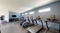 Located in Pleasant Hill, Iowa, this dedicated fitness center offers a thoughtfully designed workout space suited for a variety of exercise needs. The room features durable rubber flooring throughout, providing both safety and comfort during workouts. A row of high, horizontal windows lines one wall, allowing natural light to filter in while maintaining privacy — creating a bright yet focused atmosphere.

The cardio section is well-appointed with multiple treadmills, elliptical trainers, a stationary bike, and a rowing machine, providing ample options for aerobic exercise. On the strength training side, the space includes a multi-functional cable machine, a weight bench, a full dumbbell rack with an extensive range of free weights, medicine balls, and a plyo box for functional fitness. A wall-mounted flat-screen TV adds convenience for entertainment during workouts, while a full-length mirror supports form monitoring.

Recessed lighting in the textured ceiling ensures the space is evenly lit throughout. The cool blue-gray accent wall contrasts cleanly with the white walls and ceiling, giving the fitness center a modern, polished aesthetic. An adjacent restroom adds to the overall functionality of the space. Large windows on one end offer views of the surrounding green outdoor area, enhancing the open, energizing ambiance of this well-rounded facility.
