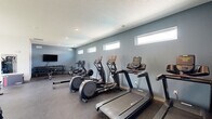 Located in Pleasant Hill, Iowa, this dedicated fitness center offers a thoughtfully designed workout space suited for a variety of exercise needs. The room features durable rubber flooring throughout, providing both safety and comfort during workouts. A row of high, horizontal windows lines one wall, allowing natural light to filter in while maintaining privacy — creating a bright yet focused atmosphere.

The cardio section is well-appointed with multiple treadmills, elliptical trainers, a stationary bike, and a rowing machine, providing ample options for aerobic exercise. On the strength training side, the space includes a multi-functional cable machine, a weight bench, a full dumbbell rack with an extensive range of free weights, medicine balls, and a plyo box for functional fitness. A wall-mounted flat-screen TV adds convenience for entertainment during workouts, while a full-length mirror supports form monitoring.

Recessed lighting in the textured ceiling ensures the space is evenly lit throughout. The cool blue-gray accent wall contrasts cleanly with the white walls and ceiling, giving the fitness center a modern, polished aesthetic. An adjacent restroom adds to the overall functionality of the space. Large windows on one end offer views of the surrounding green outdoor area, enhancing the open, energizing ambiance of this well-rounded facility.