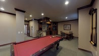 This single-level entertainment space is thoughtfully designed for leisure and hosting, combining classic styling with functional amenities. The centerpiece of the room is a full-size billiard table with a rich red felt surface and ornate dark wood legs, positioned on plush wall-to-wall carpeting that extends throughout the space. Decorative columns with dark wood crown detailing frame the open layout, lending a sense of architectural structure and elegance.

A built-in wet bar anchors one side of the room, featuring dark stained cabinetry, granite countertops, a double black basin sink, a dishwasher, and integrated diamond-pattern wine racks — both overhead and beside the counter. Bar-height wrought iron seating lines the bar's exterior, creating an inviting spot for guests. Upper cabinetry with glass displays and a built-in media niche add additional storage and entertainment functionality.

A dedicated dining area with a wooden table and upholstered chairs offers a comfortable space for more relaxed gatherings. Framed artwork decorates the neutral-toned walls, while recessed lighting throughout provides warm, even illumination. Windows fitted with wood-slat blinds allow for natural light, and a large framed mirror enhances the sense of depth and openness. Staircase access is visible, connecting the space to the rest of the home.