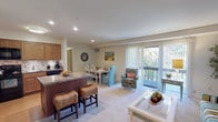 This single-floor residence offers a thoughtfully designed layout that balances comfort and functionality. The open-concept living and dining area serves as the heart of the home, filled with natural light streaming through large sliding glass doors that lead to a private balcony overlooking lush greenery. The living room features plush carpeting and generous space for seating, making it ideal for both relaxing and entertaining.

Adjacent to the living area, the dining space comfortably accommodates a full-sized dining table, seamlessly connecting to the kitchen. The kitchen is well-equipped with warm wood cabinetry, granite-style countertops, a mosaic tile backsplash, and black appliances including a range, microwave, refrigerator, and dishwasher. A peninsula with bar seating adds both prep space and a casual dining option.

The residence includes multiple bedrooms, each carpeted and featuring ample closet storage with louvered bifold doors. The primary bedroom is a quiet retreat with room for a full bedroom suite, while a secondary bedroom offers versatility with generous closet space. The bathroom is clean and functional, featuring a soaking tub, tiled flooring, and a vanity with storage. Throughout the home, neutral tones and recessed lighting create a bright, welcoming ambiance.