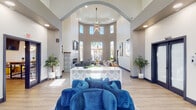 This single-floor community clubhouse is a thoughtfully designed gathering space that blends contemporary style with welcoming comfort. The centerpiece of the space is a dramatic rotunda-style lobby featuring soaring double-height ceilings, a grand archway, and a statement sputnik chandelier that draws the eye upward. Rows of tall, narrow windows bathe the space in natural light while framing views of palm trees and the surrounding outdoor area. A central reception desk with an intricate geometric tile façade anchors the main lounge, which is furnished with plush, jewel-toned velvet seating arranged for casual conversation.

A fireplace clad in bold black-and-white patterned encaustic tile serves as a striking focal point in one of the lounge alcoves, complemented by curated artwork, metallic wall sculptures, and accent mirrors. Tall floor-to-ceiling slit windows throughout add architectural drama while maintaining a connection to the lush outdoor landscaping. The warm-toned luxury vinyl plank flooring runs consistently throughout, unifying the various zones of the space.

Adjacent to the main lounge is a dedicated co-working and gathering area featuring a large communal wood-top table, mixed seating, pendant lighting, a wall-mounted television, and vibrant accent furnishings. This multifunctional room is ideal for both collaborative work and casual socializing, completing this well-appointed community amenity space.